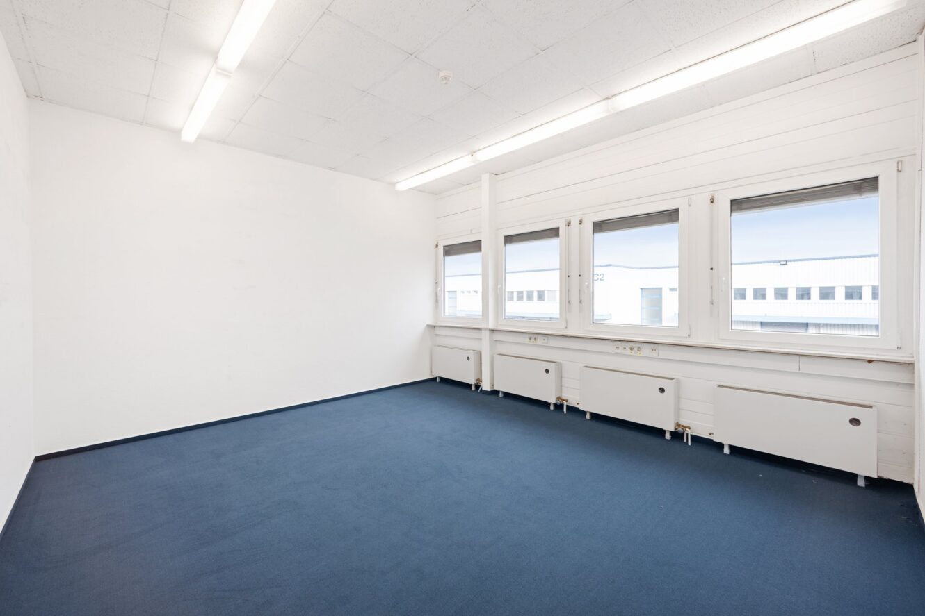 Leerer Büroraum mit blauem Teppich, weißen Wänden und Decken, Leuchtstoffröhren und drei großen Fenstern mit Heizkörpern darunter.