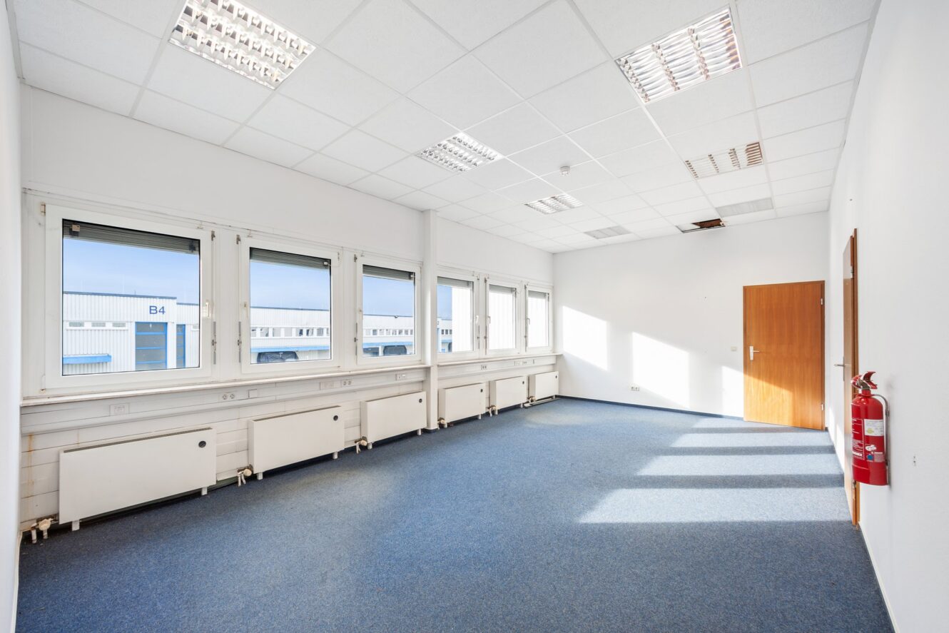 Leerer Büroraum mit blauem Teppich, weißen Wänden, großen Fenstern, fluoreszierenden Deckenleuchten, einer geschlossenen Holztür und einem roten Feuerlöscher an der Wand.