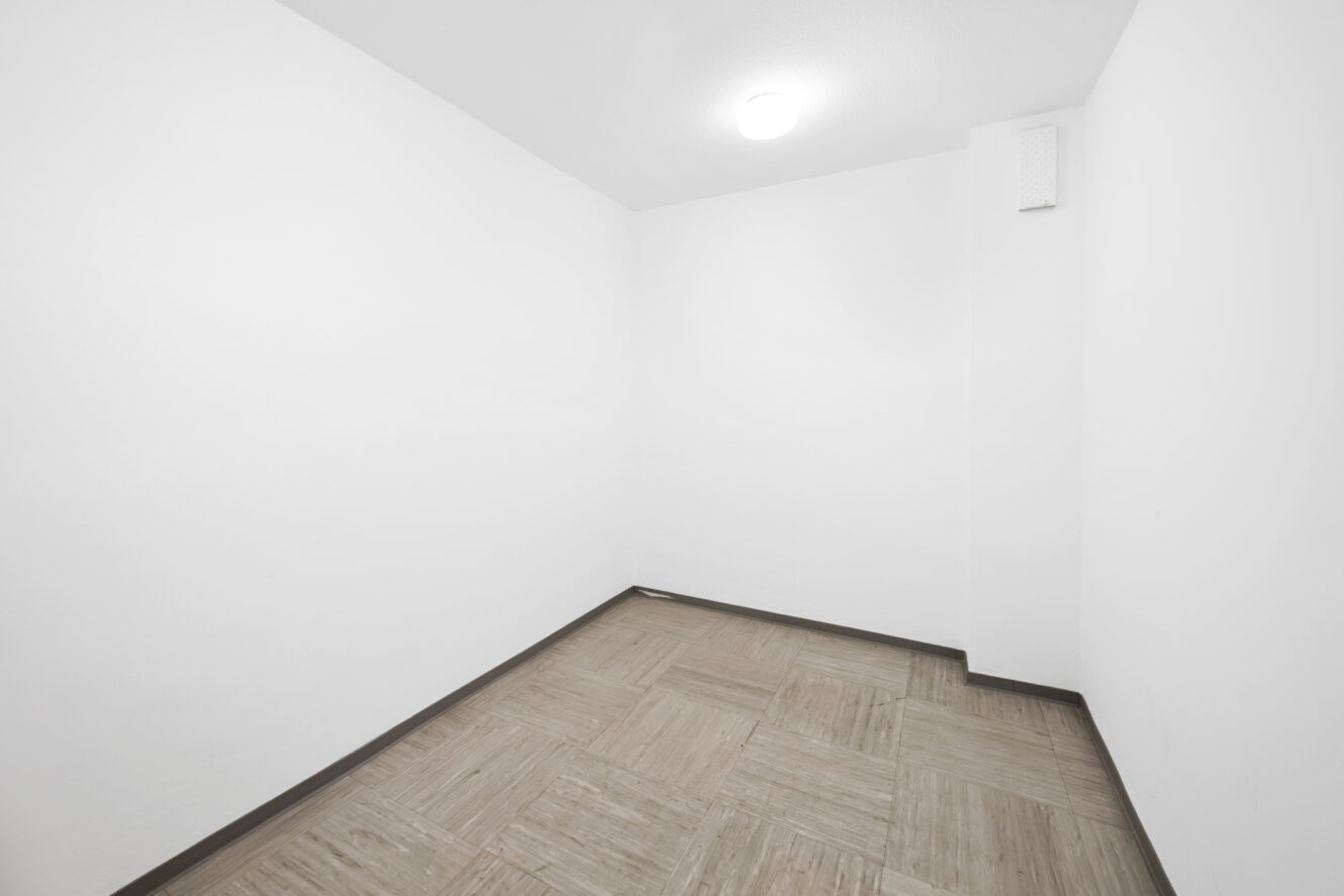 Kleiner, leerer Raum mit weißen Wänden, einer Leuchte an der Decke und hellem Kachelboden.