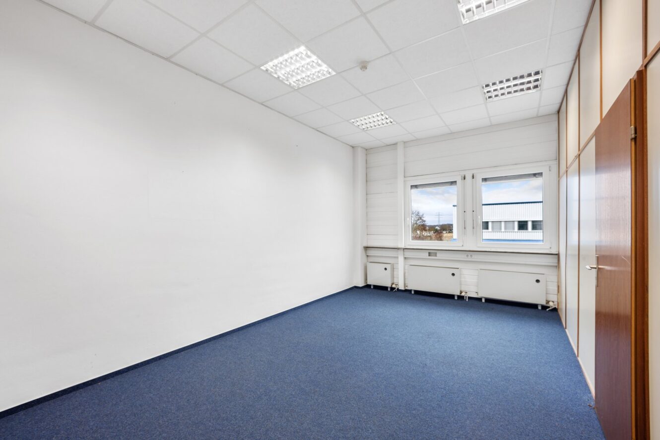 Leerer Büroraum mit weißen Wänden, blauem Teppich, Deckenleuchten, zwei Fenstern und einer Holztür auf der rechten Seite.