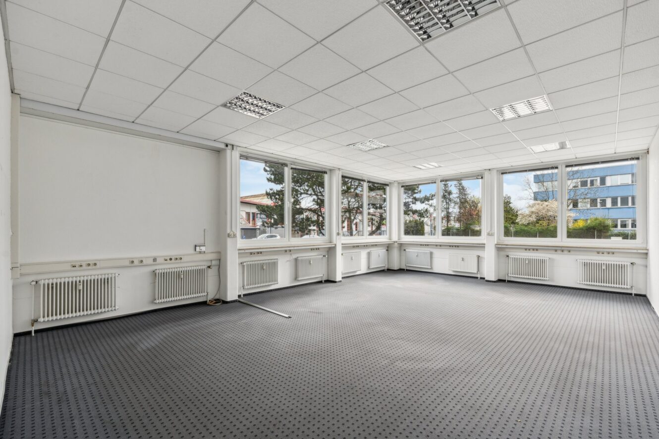 Ein leerer Büroraum mit großen Fenstern, weißen Wänden, einer Gitterdecke mit Leuchtstoffröhren, Heizkörpern und einem dunkel gesprenkelten Boden.