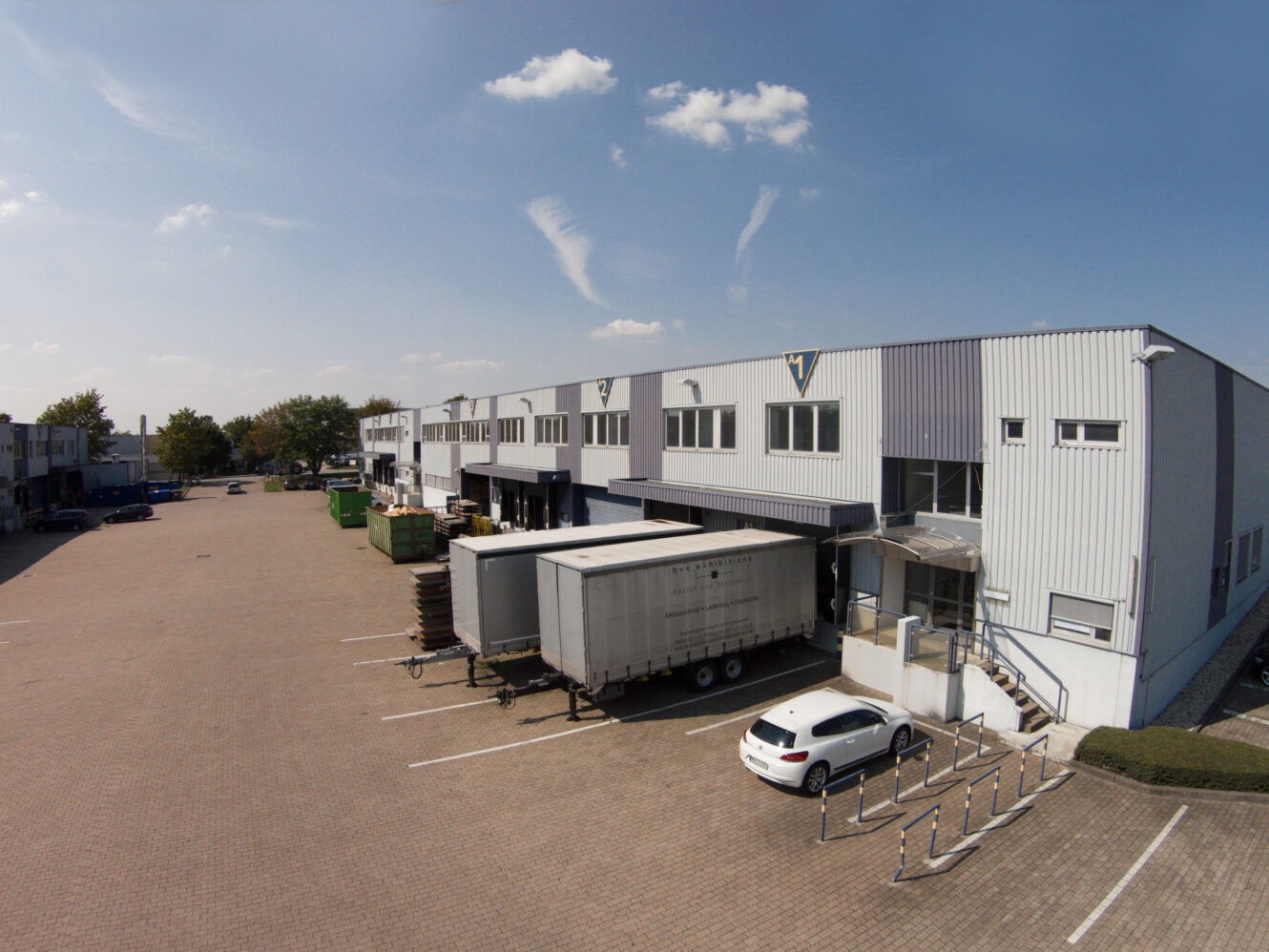 Ein großes Industrielager mit Laderampen, ein weißer Lkw-Anhänger auf dem Rücksitz und ein weißes Auto, das auf einem größtenteils leeren, gepflasterten Grundstück unter freiem Himmel geparkt ist.