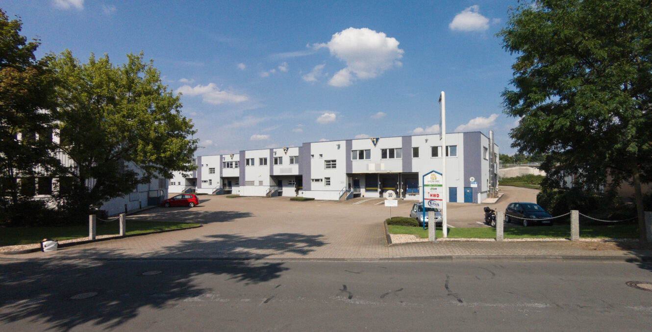 Eine Reihe moderner, weißer Industriegebäude mit mehreren Garagentoren und Parkplätzen, umgeben von Bäumen und Autos, unter einem teilweise bewölkten Himmel.