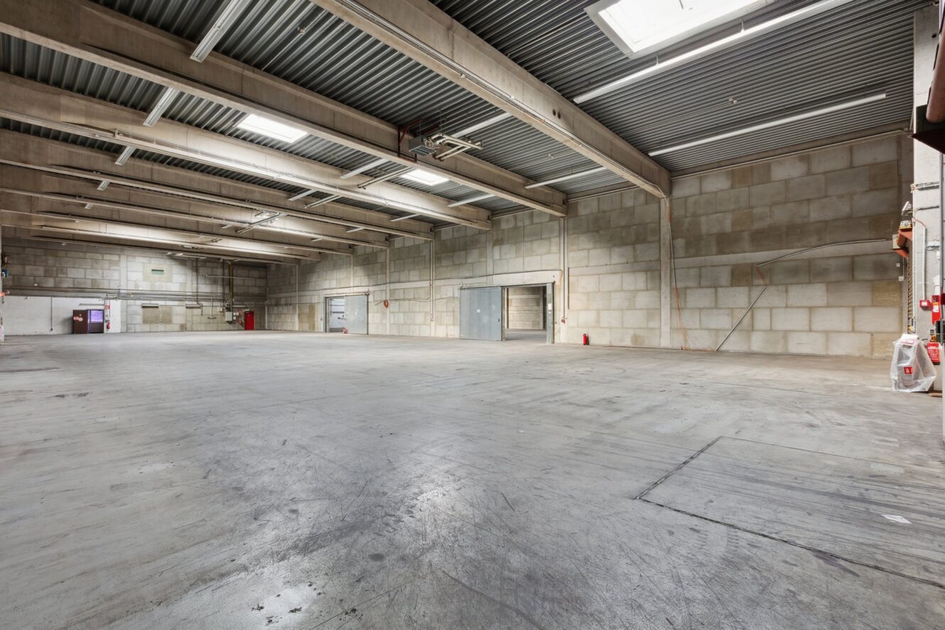 Große, leere Lagerhalle mit Betonböden, hohen Decken, freiliegenden Balken und Leuchtstoffröhrenbeleuchtung. An den Wänden sind mehrere Türen und Feuerlöscher zu sehen.
