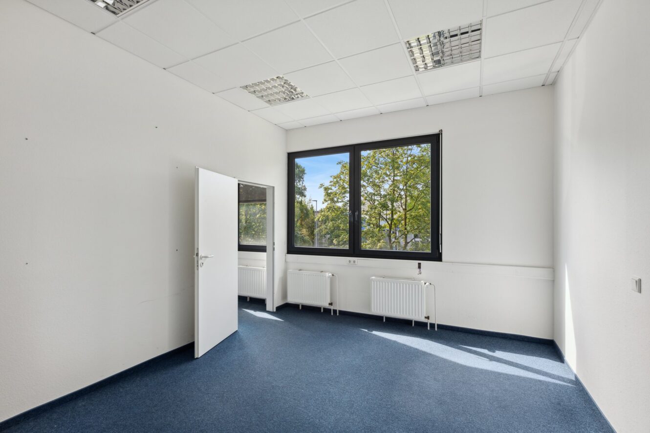 Leerer Büroraum mit weißen Wänden, blauem Teppich, einer offenen Tür, zwei großen Fenstern und Heizkörpern unter den Fenstern. Draußen sind Bäume zu sehen.