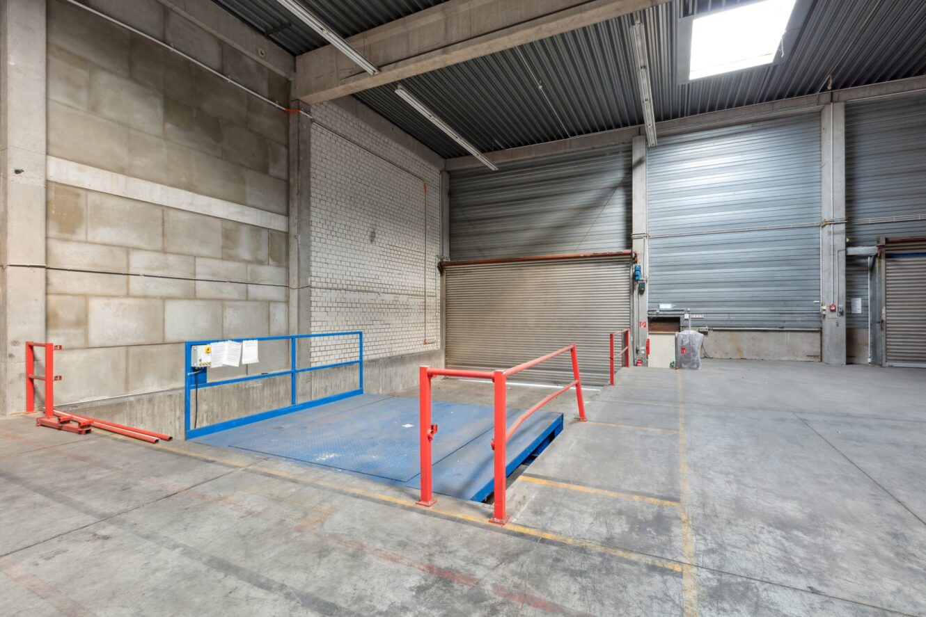 Eine Laderampe mit blauer Metallplattform und rotem Geländer in einer leeren Industriehalle mit Betonwänden und geschlossenen Rolltoren.
