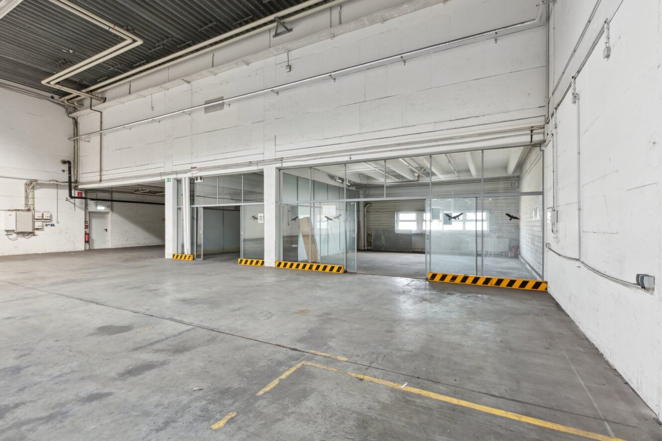 Großes, leeres Industrielager mit hohen Decken, Betonböden und verglasten Büroräumen an einer Seite. Gelbe und schwarze Stoßstangen säumen die Eingänge zu den Büros.