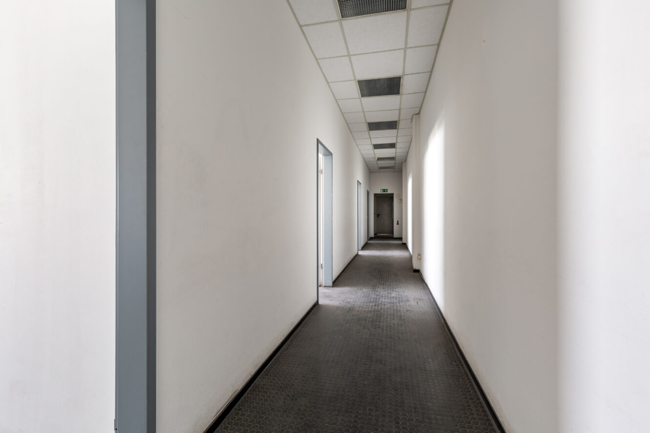 Ein langer, leerer Flur mit weißen Wänden, geschlossenen Türen auf beiden Seiten, einer gekachelten Decke und natürlichem Licht, das durch die Fenster auf der rechten Seite fällt.