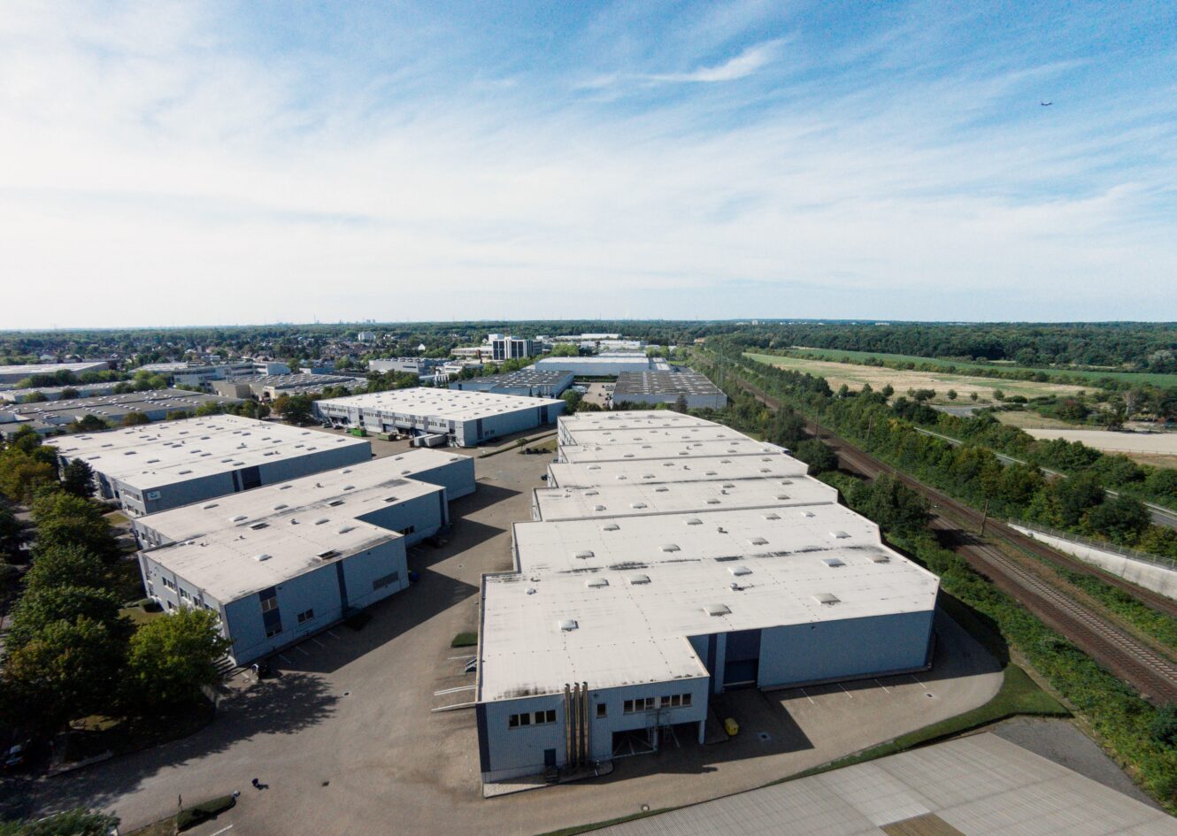 Luftaufnahme eines großen Industriekomplexes mit mehreren Lagergebäuden, umgeben von Bäumen und Feldern, neben einer Eisenbahnlinie bei teilweise bewölktem Himmel.