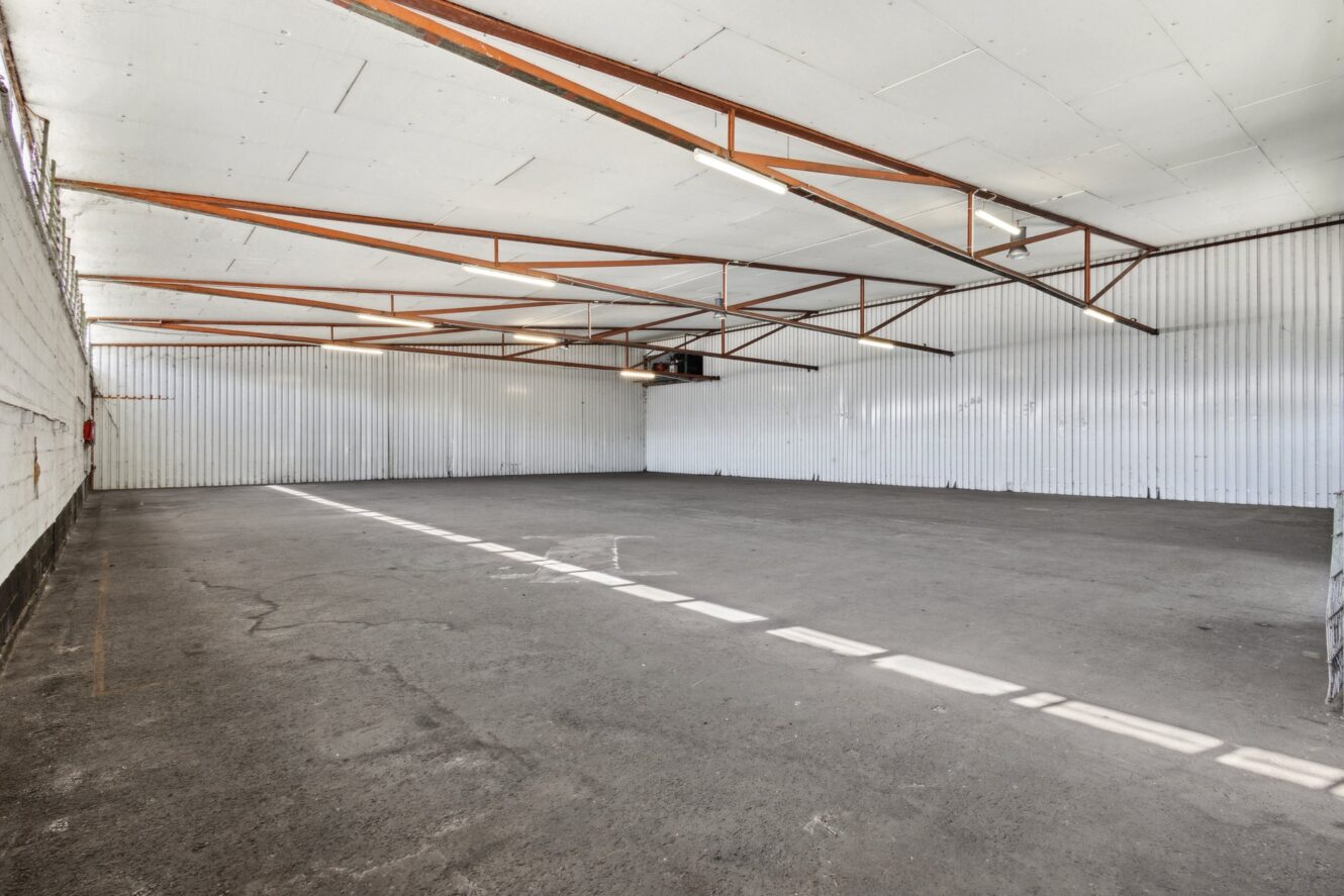 Große leere Lagerhalle mit weißen Wellblechwänden, Betonboden, freiliegenden Stahldachträgern und Leuchtstoffröhren. Sonnenlicht strömt durch die Fenster an der linken Wand.