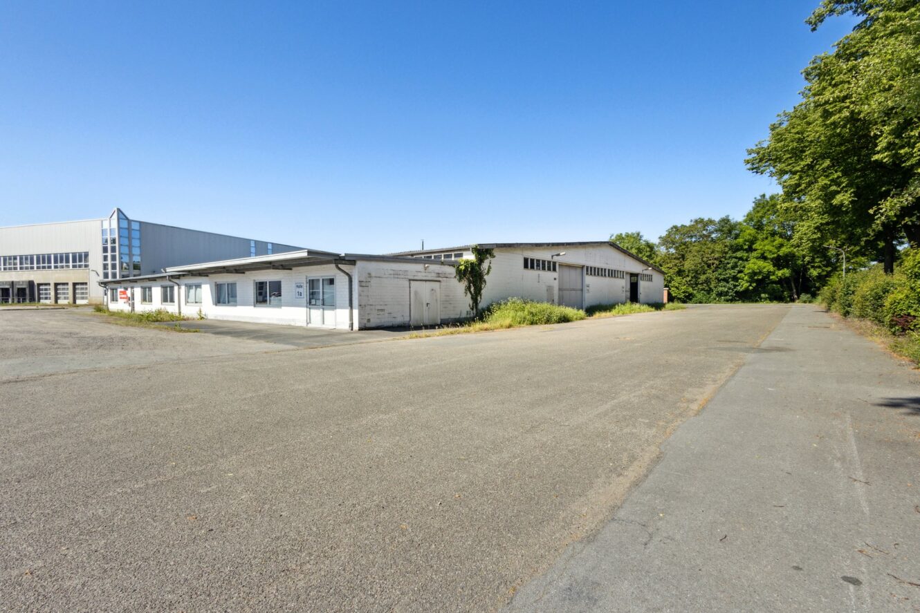 Ein großer, leerer, gepflasterter Platz mit einem weißen Industriegebäude auf der linken Seite; Bäume und Grünflächen säumen den rechten Rand unter einem klaren, blauen Himmel.