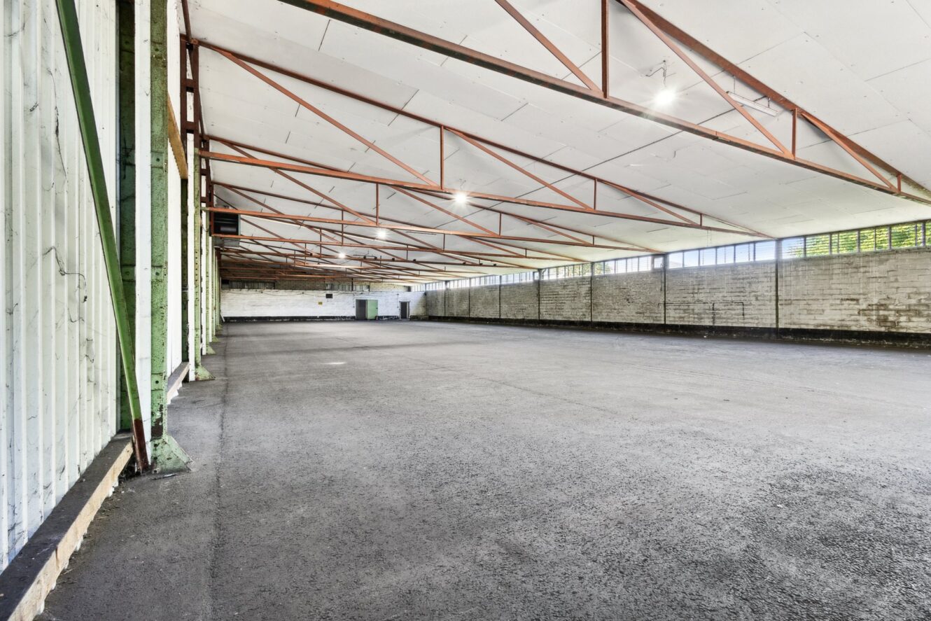 Innenansicht einer großen, leeren Lagerhalle mit freiliegenden Metallträgern, weißen Deckenplatten, Betonwänden und einem nackten Betonboden. Natürliches Licht fällt durch hohe horizontale Fenster ein.