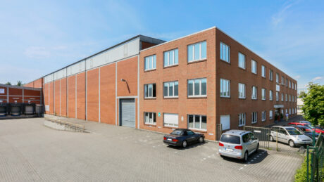 Ein großes, dreistöckiges Lagerhaus aus Backstein mit mehreren Fenstern, einer Laderampe und mehreren geparkten Autos auf dem angrenzenden Parkplatz an einem klaren Tag.