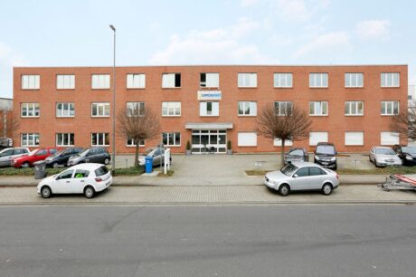 Ein dreistöckiges Bürogebäude aus Backstein mit mehreren Fenstern, einem zentralen Eingang, geparkten Autos davor und einem Schild über der Tür mit der Aufschrift "comspace".