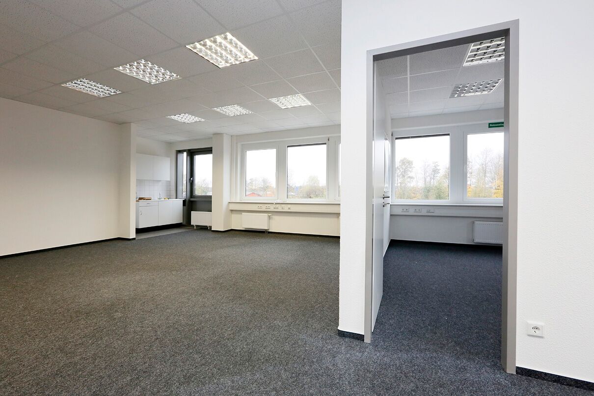 Ein leerstehender Büroraum mit grauem Teppich, weißen Wänden, fluoreszierenden Deckenleuchten, großen Fenstern und einem angrenzenden kleinen Raum, der durch eine offene Tür zu sehen ist.