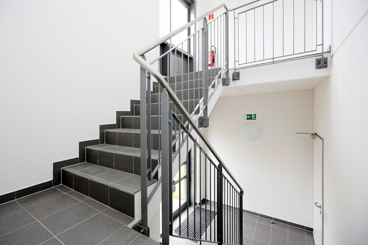 Moderne Innentreppe mit dunkelgrauen Fliesen und Metallgeländer, die zu einem oberen Treppenabsatz mit einem Feuerlöscher und einem Notausgangsschild an der Wand führt.