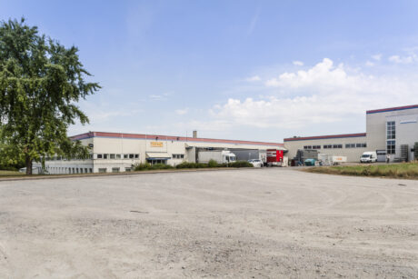Große Industriehalle mit weißem und rotem Äußeren, mehrere in der Nähe geparkte Lastwagen und Lieferwagen und ein leerer Kiesplatz im Vordergrund unter blauem Himmel.