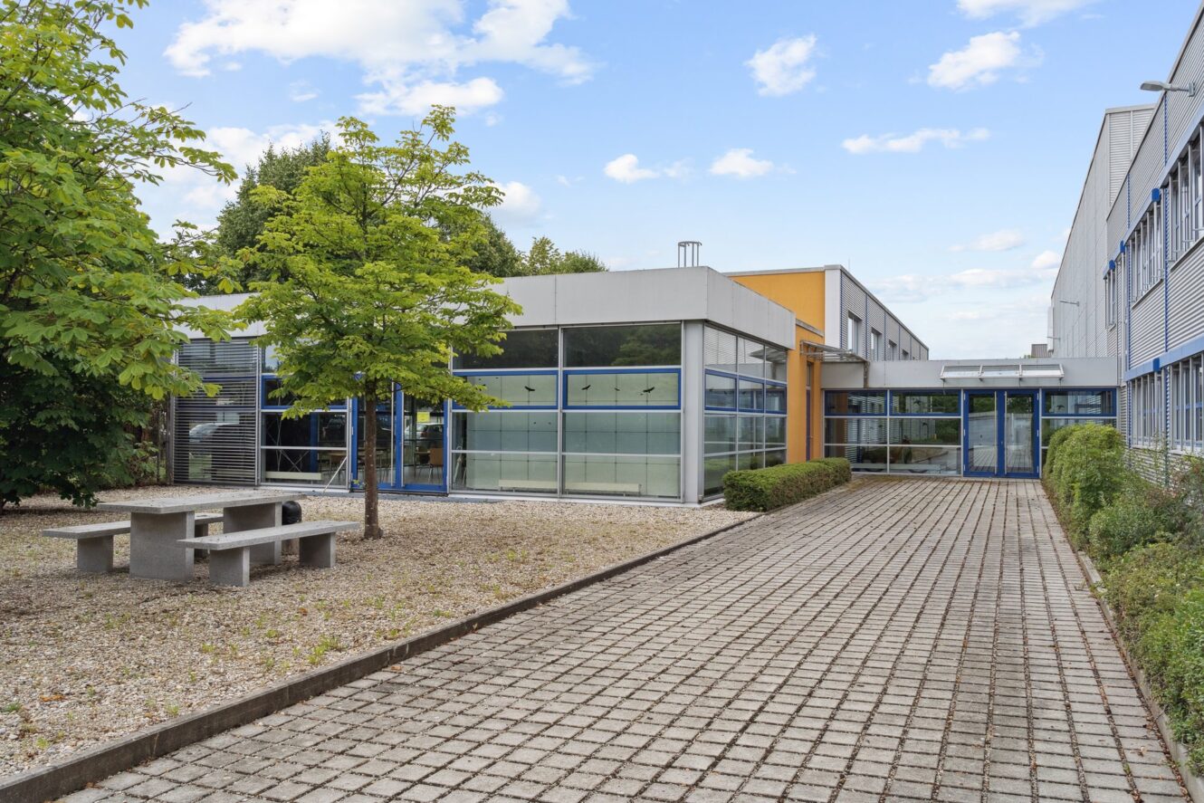 Ein modernes Gebäude mit großen Fenstern, einem gepflasterten Gehweg, einer Kiesfläche, einem Picknicktisch aus Beton und grünen Bäumen unter einem teilweise bewölkten Himmel.