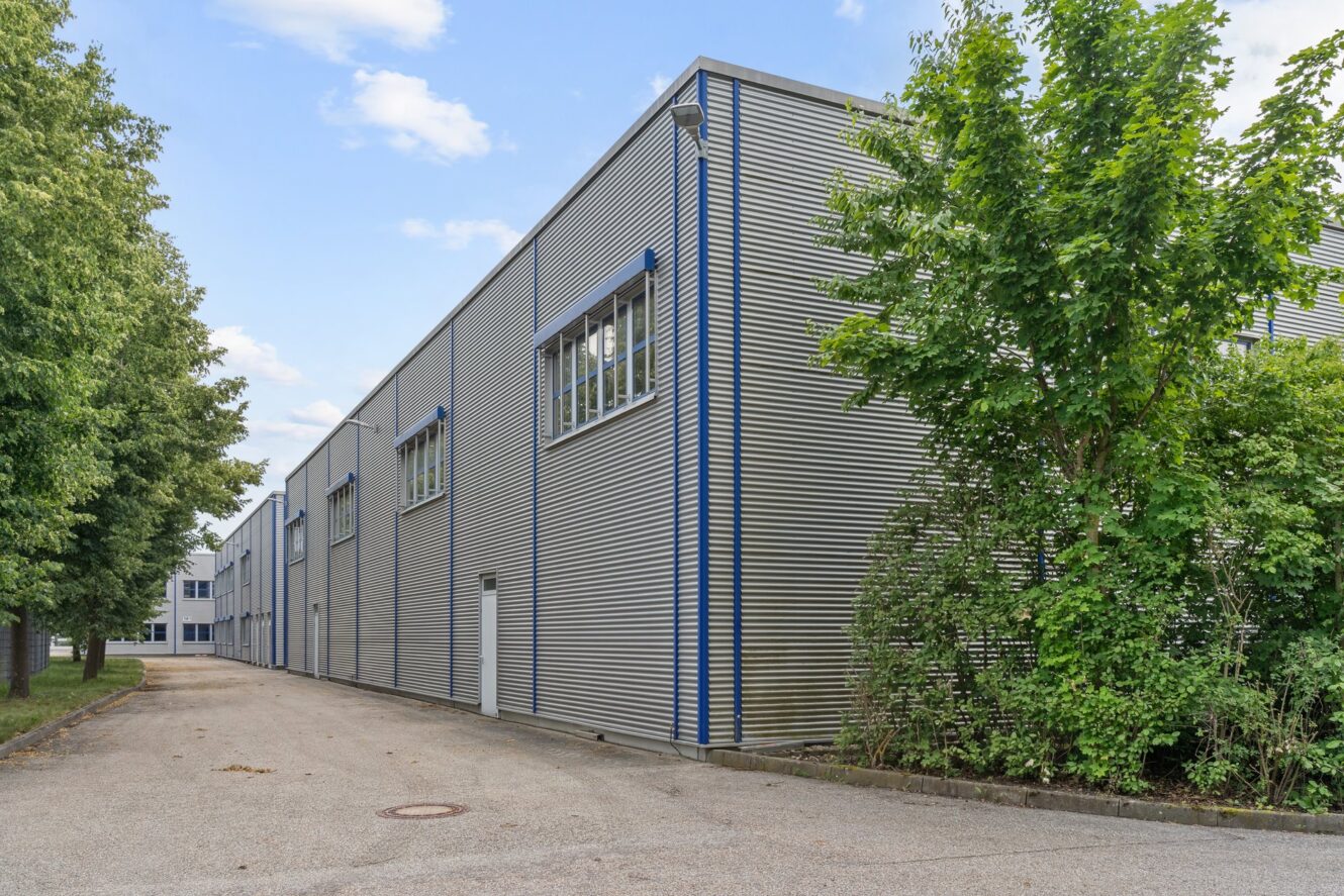 Ein metallenes Industriegebäude mit großen Fenstern und blauen Verzierungen steht neben einer gepflasterten Auffahrt, gesäumt von grünen Bäumen und Büschen unter einem teilweise bewölkten Himmel.