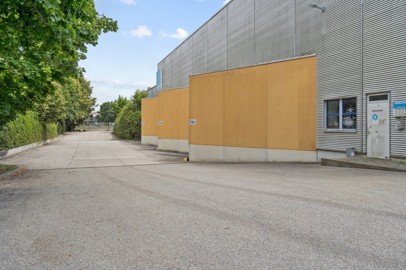 Ein Industriegebäude mit gelben Wänden, einer eingezäunten Auffahrt, Bäumen auf der linken Seite und einem klaren Himmel darüber.