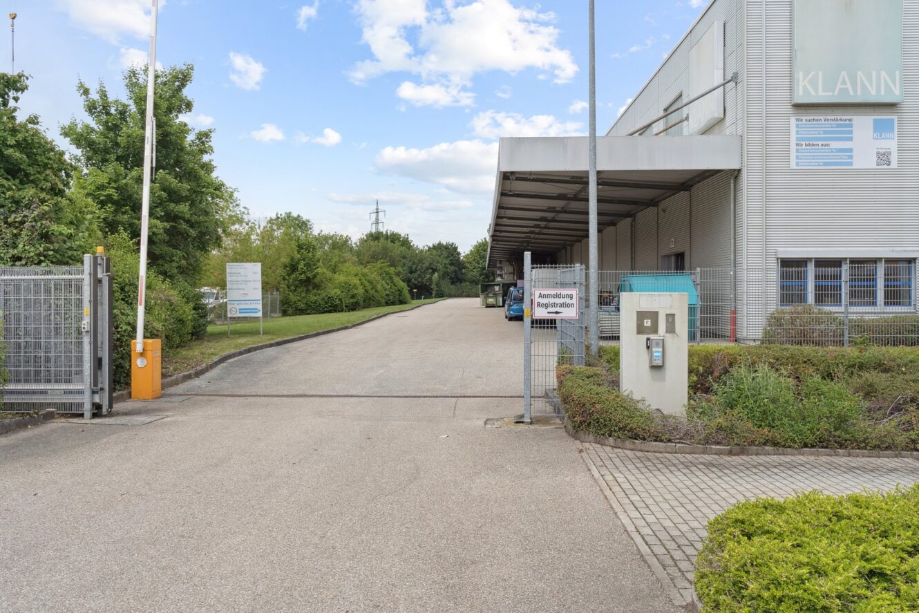 Einfahrt zu einer Industrieanlage mit einem Sicherheitstor, Schildern und einem großen Gebäude mit der Aufschrift KLANN auf der rechten Seite, umgeben von Grünflächen unter einem teilweise bewölkten Himmel.
