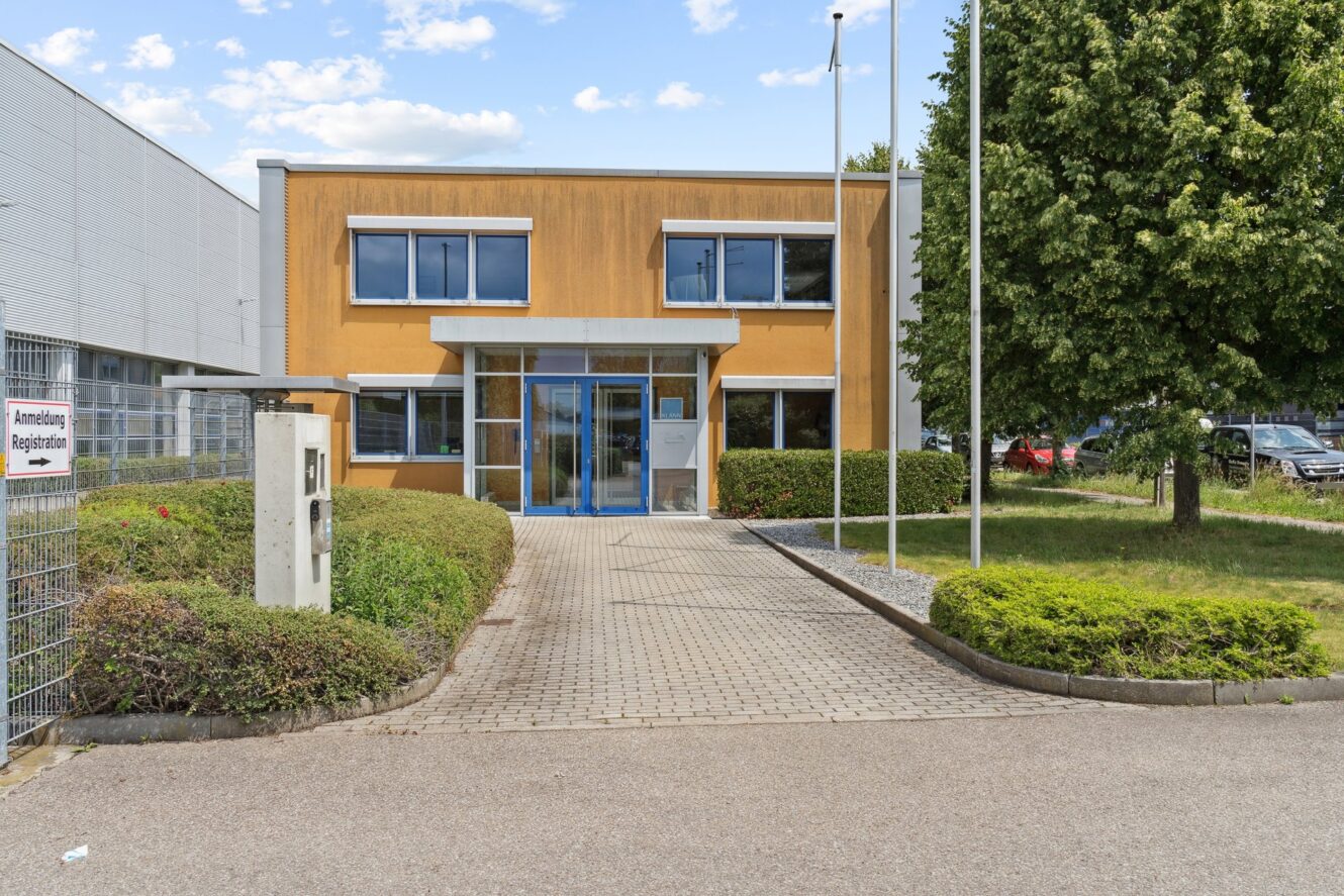 Ein zweistöckiges orangefarbenes Gebäude mit blau gerahmten Fenstern und Glastüren, umgeben von Grün und einem gepflasterten Gehweg, der zum Eingang führt.