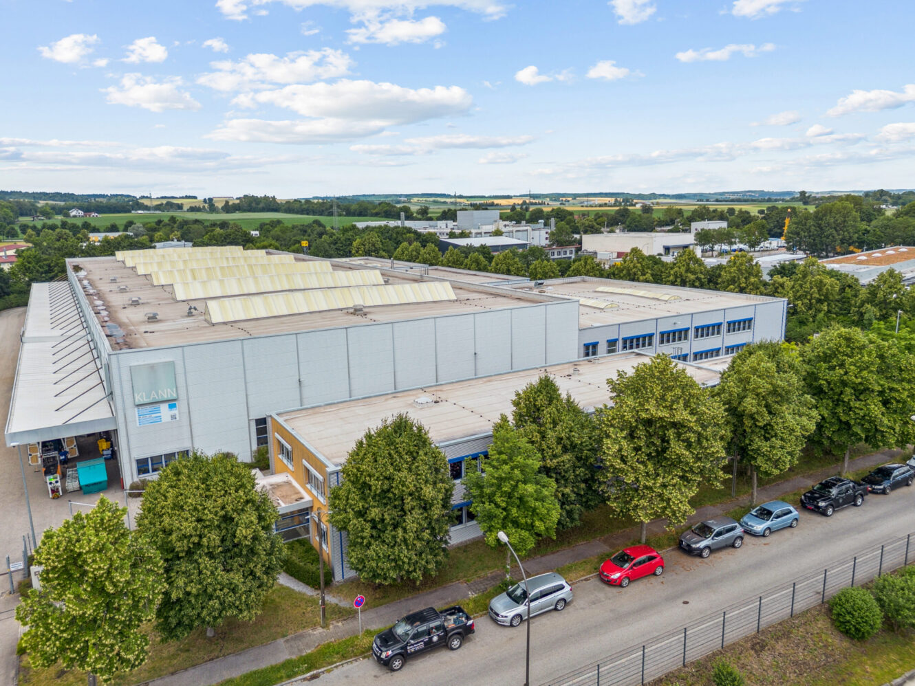 Luftaufnahme eines großen Industrielagers mit einem blauen Schild, umgeben von Bäumen, geparkten Autos und einer ländlichen Landschaft im Hintergrund.