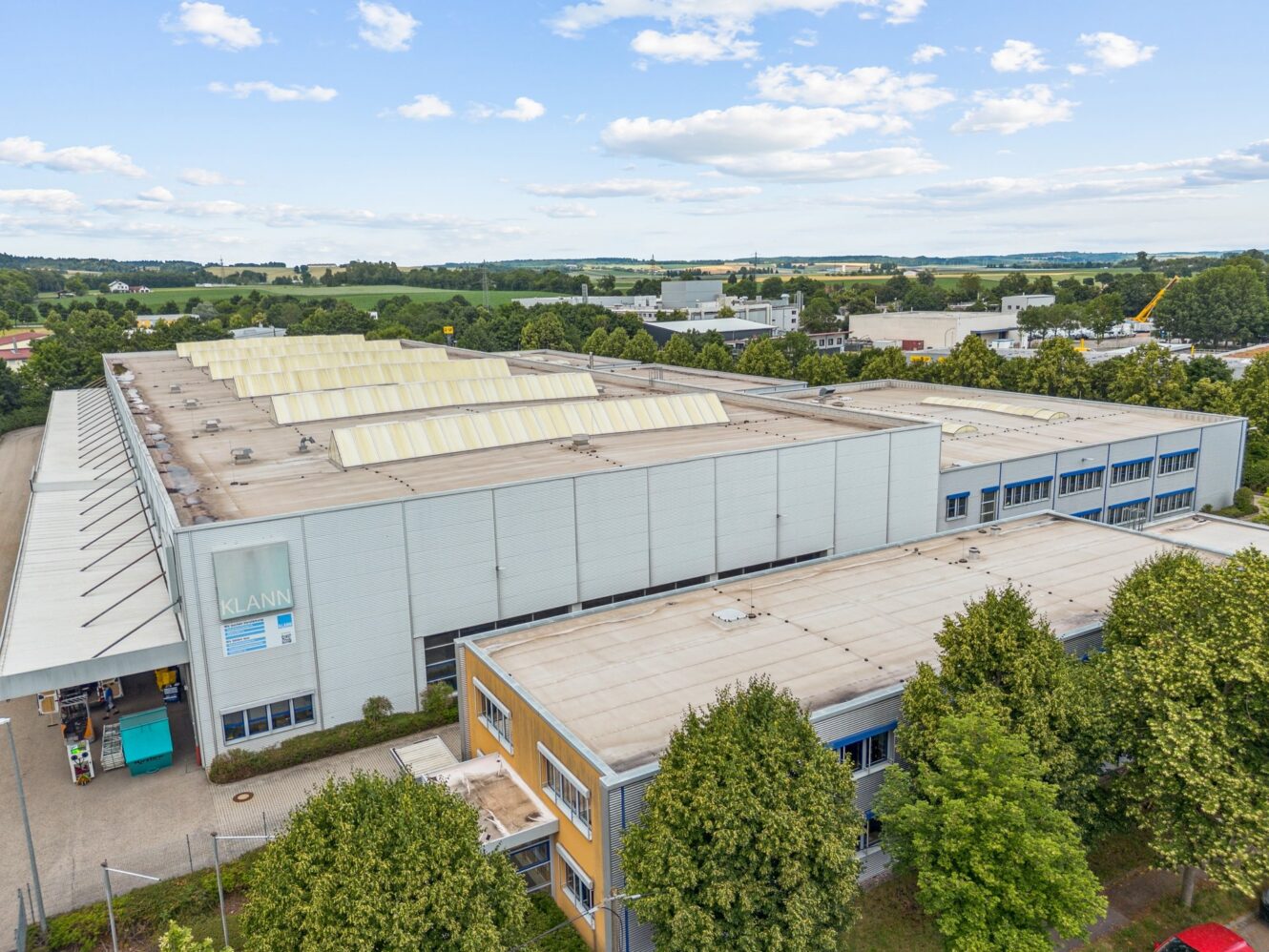 Ein großes industrielles Lagergebäude mit Flachdach und Oberlichtern, umgeben von Bäumen und angrenzend an ein kleineres Bürogebäude, unter einem teilweise bewölkten Himmel.