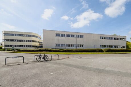 Ein modernes Bürogebäude mit Flachdach, großen Fenstern, Fahrradständern und einem Fahrrad davor, unter einem blauen Himmel mit vereinzelten Wolken.