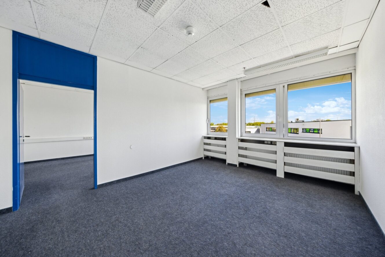 Leerer Büroraum mit weißen Wänden, blauem Teppich, Neonbeleuchtung und zwei großen Fenstern, die eine Stadtlandschaft und einen blauen Himmel zeigen. Die Deckenplatten scheinen leicht beschädigt zu sein.