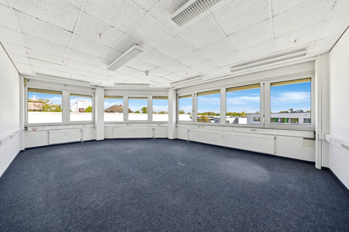 Leerer Büroraum mit blauem Teppich, weißen Wänden und großen Fenstern an zwei Seiten, die eine Stadtlandschaft im Freien mit blauem Himmel und einigen Wolken zeigen. An der Decke hängen Leuchtstoffröhren.