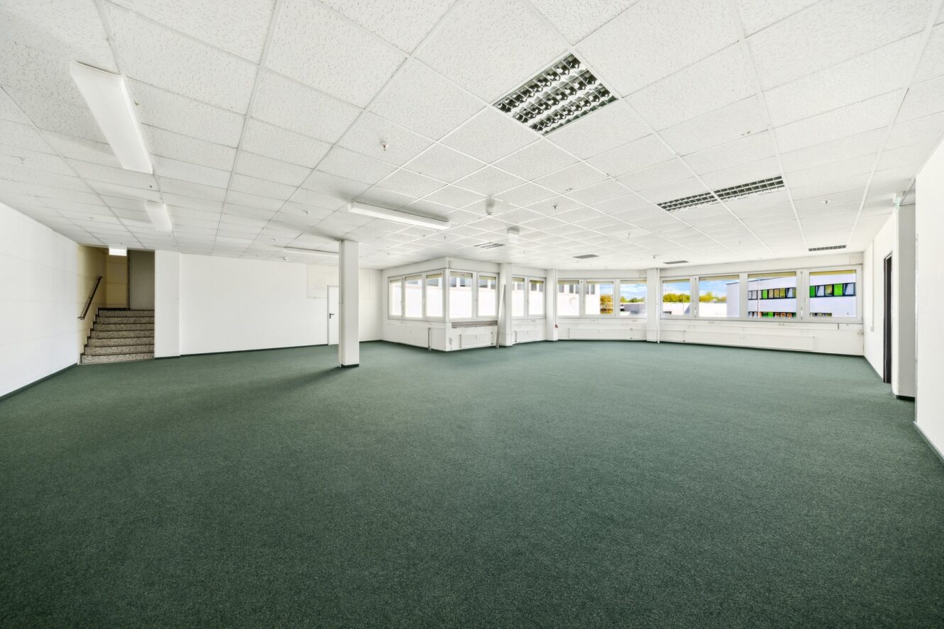 Leere Büroräume mit grünem Teppich, weißen Wänden, Neonröhren an der Decke und großen Fenstern an zwei Seiten, wobei in der hinteren Ecke eine Treppe zu sehen ist.