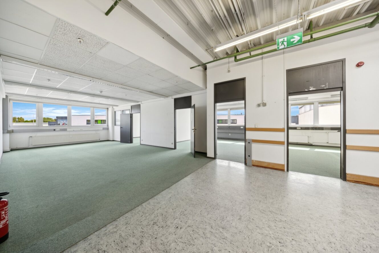 Leere Büroräume mit grünem und weißem Bodenbelag, großen Fenstern, weißen Wänden, freiliegenden Deckenrohren und sichtbaren Notausgangsschildern.