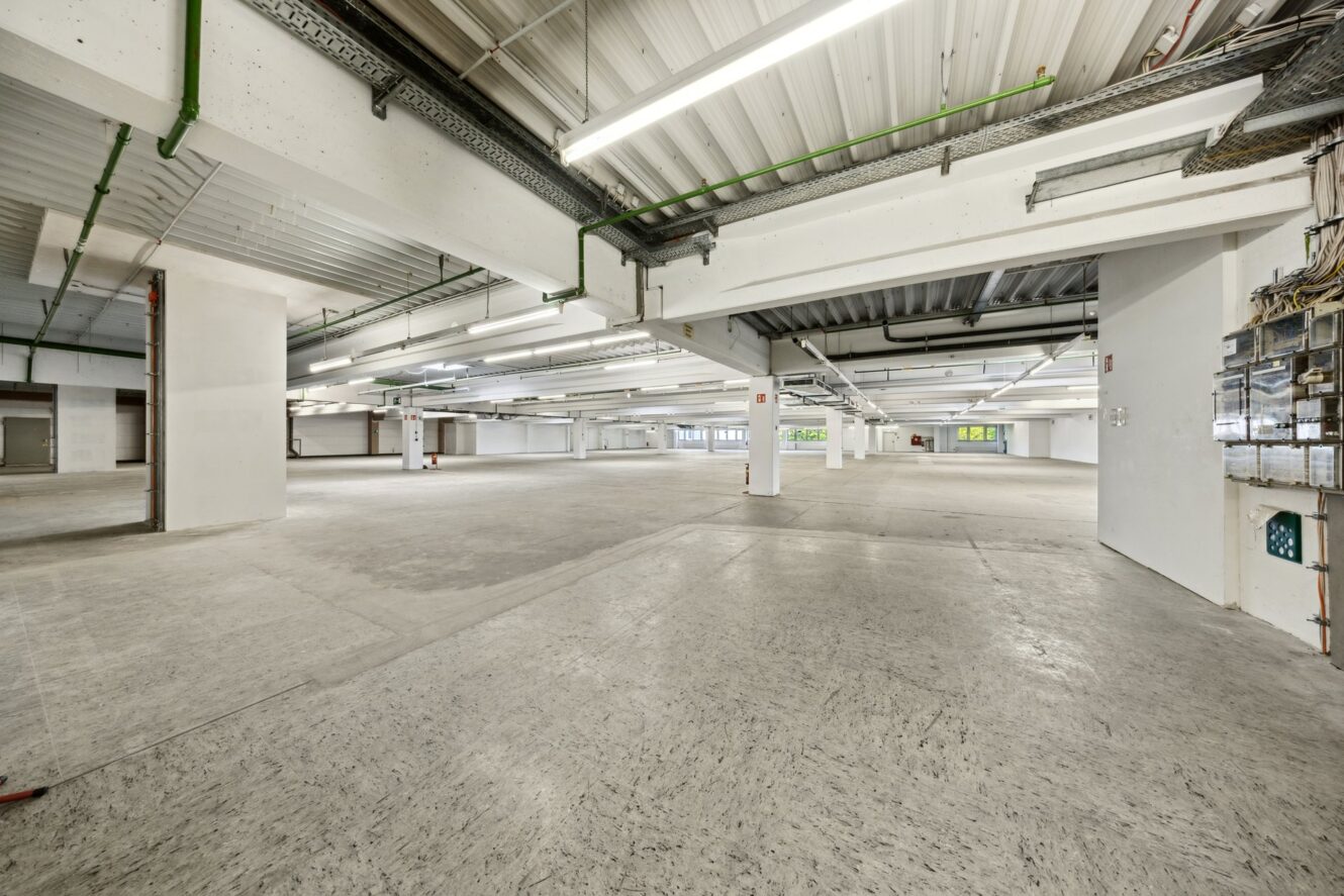 Geräumiges, leeres Industrielager mit Betonböden, weißen Säulen, fluoreszierenden Deckenleuchten und freiliegenden Rohrleitungen.