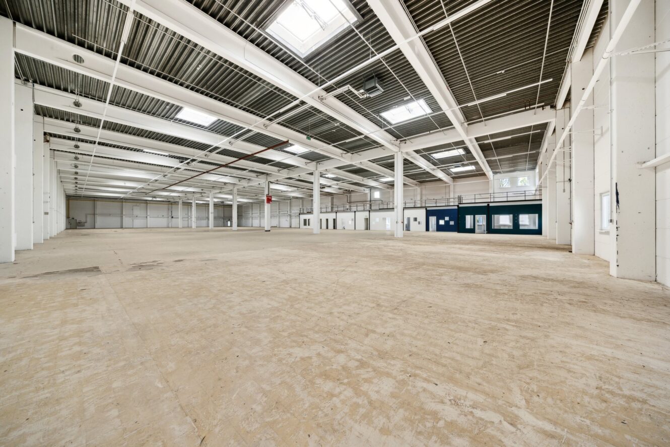 Großes leeres Industrielager mit hohen Decken, sichtbaren Balken, Oberlichtern und einem kleinen Bürobereich an einer Wand.