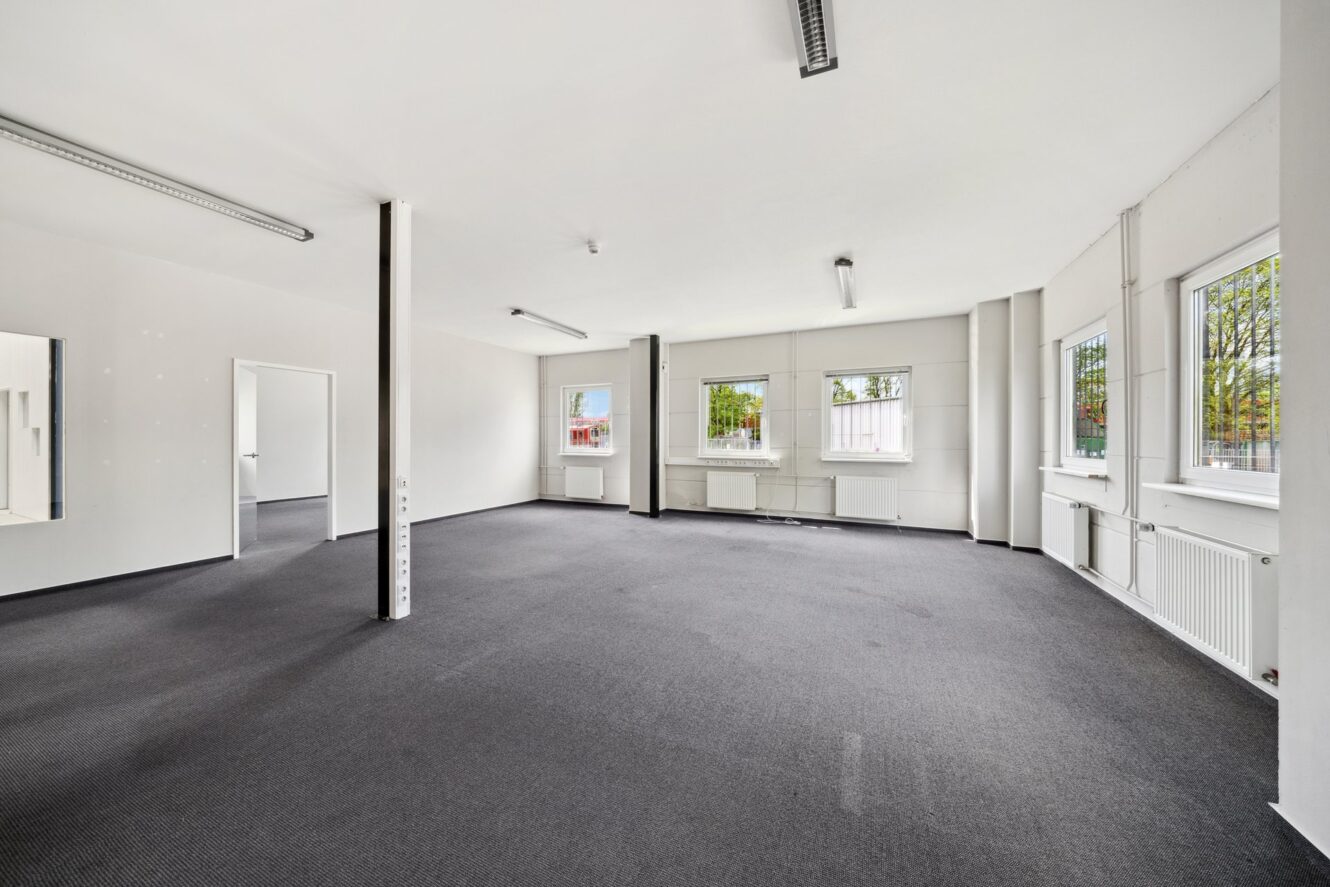 Leere Büroräume mit weißen Wänden, grauem Teppich, fluoreszierenden Deckenleuchten, mehreren Fenstern und ein paar Wandsäulen.