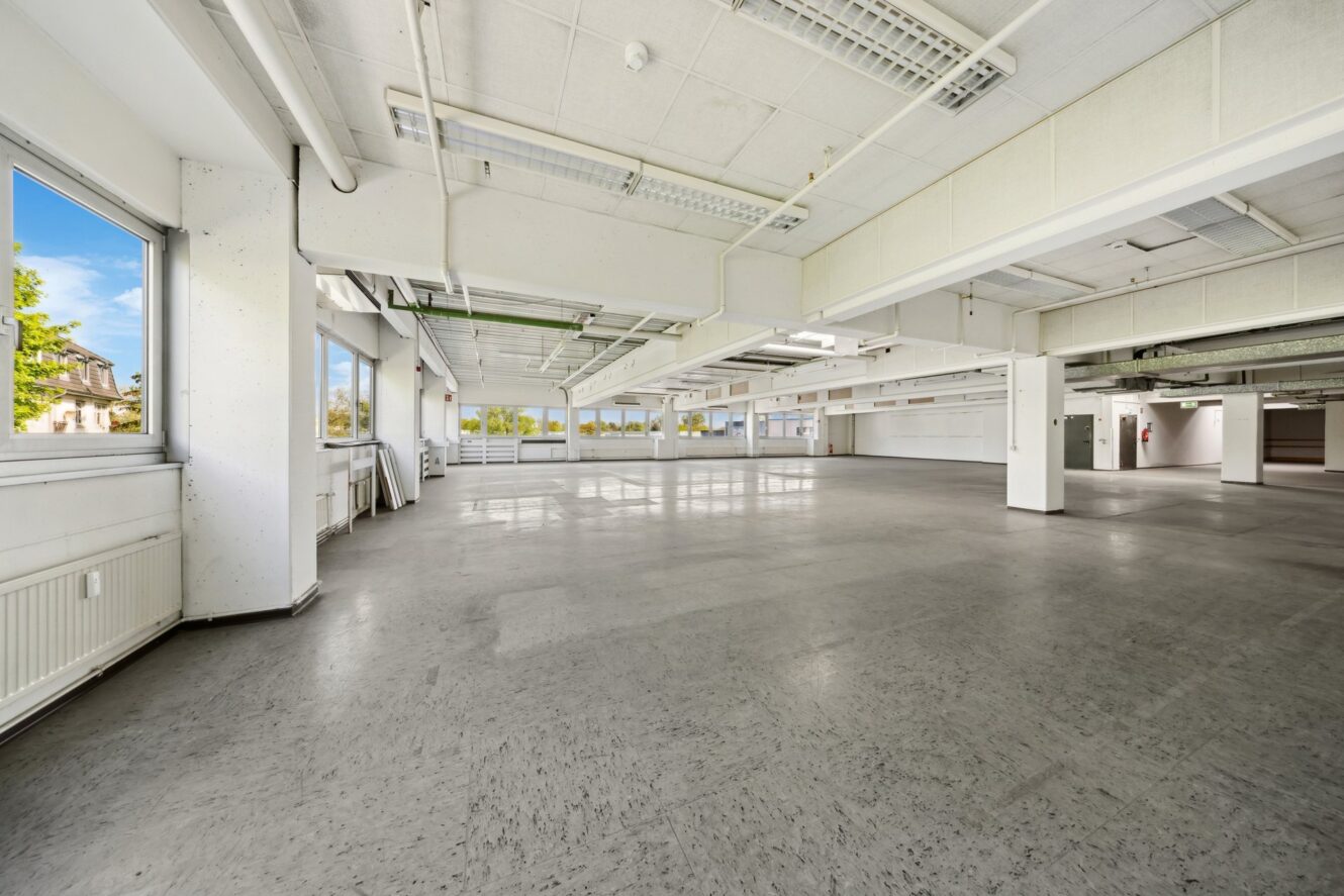 Geräumiger, leerer Raum im Industriestil mit großen Fenstern, weißen Wänden, Deckenbeleuchtung und grauem Bodenbelag.