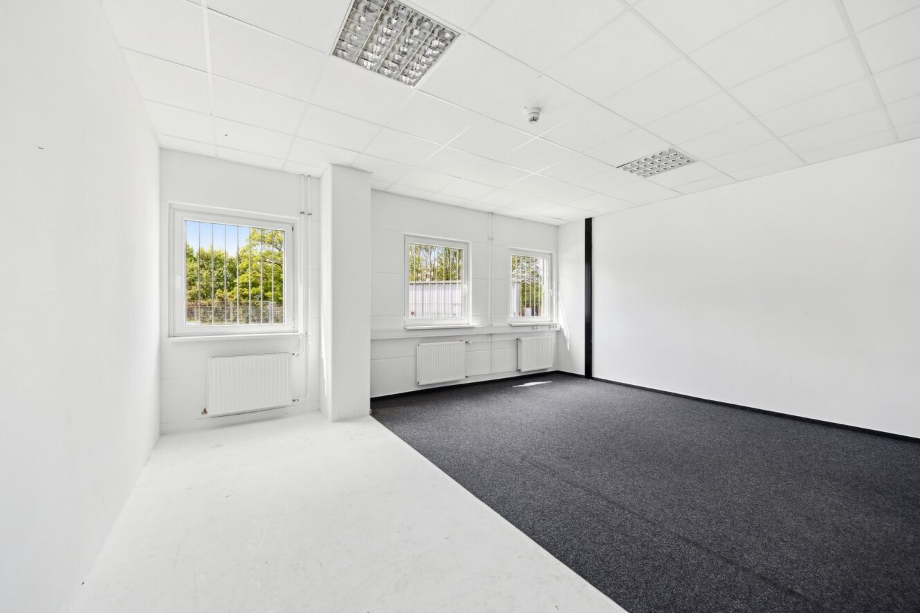 Leere, unmöblierte Büroräume mit weißen Wänden, vergitterten Fenstern, einer Mischung aus weißen Kacheln und schwarzem Teppichboden und fluoreszierenden Deckenleuchten.