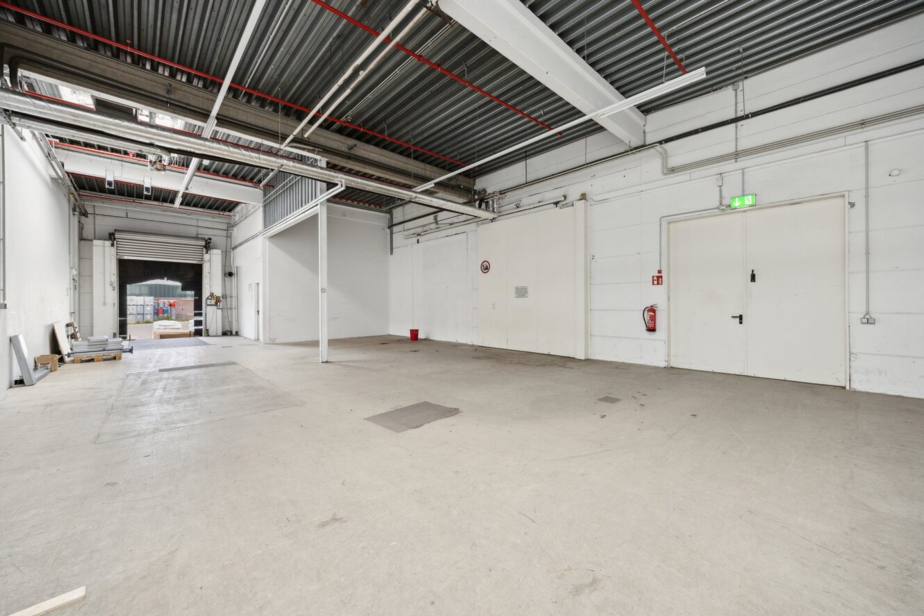 Großer, leerer Industrielagerraum mit Betonboden, weißen Wänden, hoher Decke, freiliegenden Rohren, Feuerlöscher und einer großen offenen Tür, die nach draußen führt.