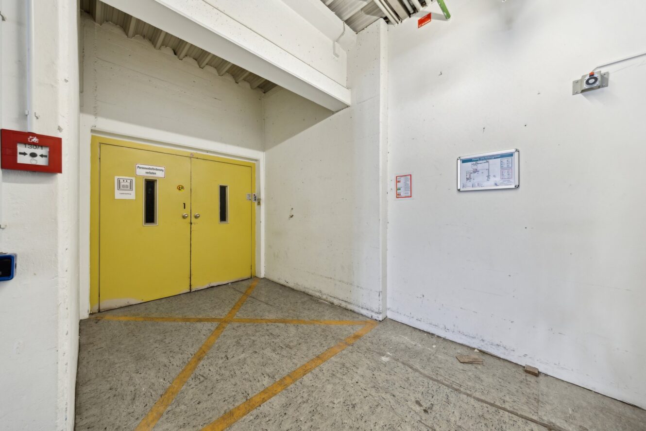 Industriehalle mit gelben Doppeltüren, weißen Wänden, Sicherheitsschildern, einer an der Wand montierten Kamera und einem mit gelben diagonalen Linien markierten Boden.