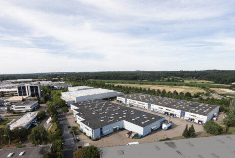 Luftaufnahme eines Industriekomplexes mit großen Lagergebäuden, Parkplätzen und umliegenden Grünflächen bei teilweise bewölktem Himmel.