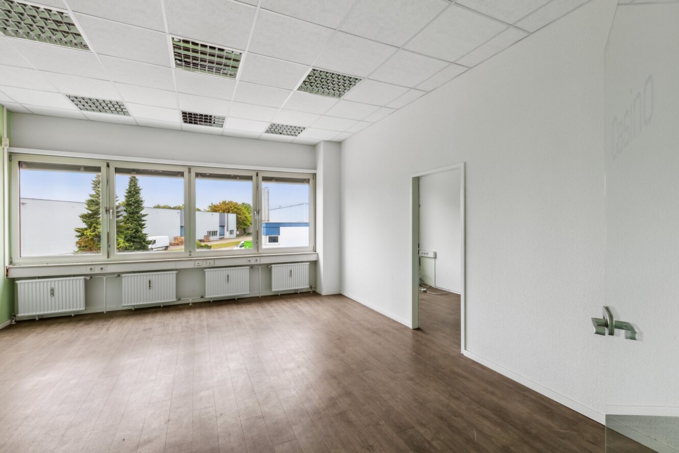 Leerer Büroraum mit weißen Wänden, großen Fenstern, braunem Holzfußboden und Deckenleuchten; ein kleineres angrenzendes Büro ist durch eine offene Tür sichtbar.