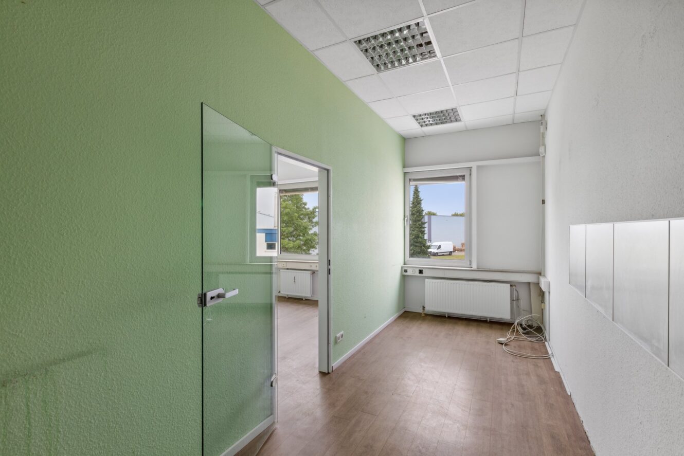 Ein schmales, leeres Büro mit grünen und weißen Wänden, Holzfußboden, einer Glastür und einem Fenster mit Blick auf einen Parkplatz und Bäume.