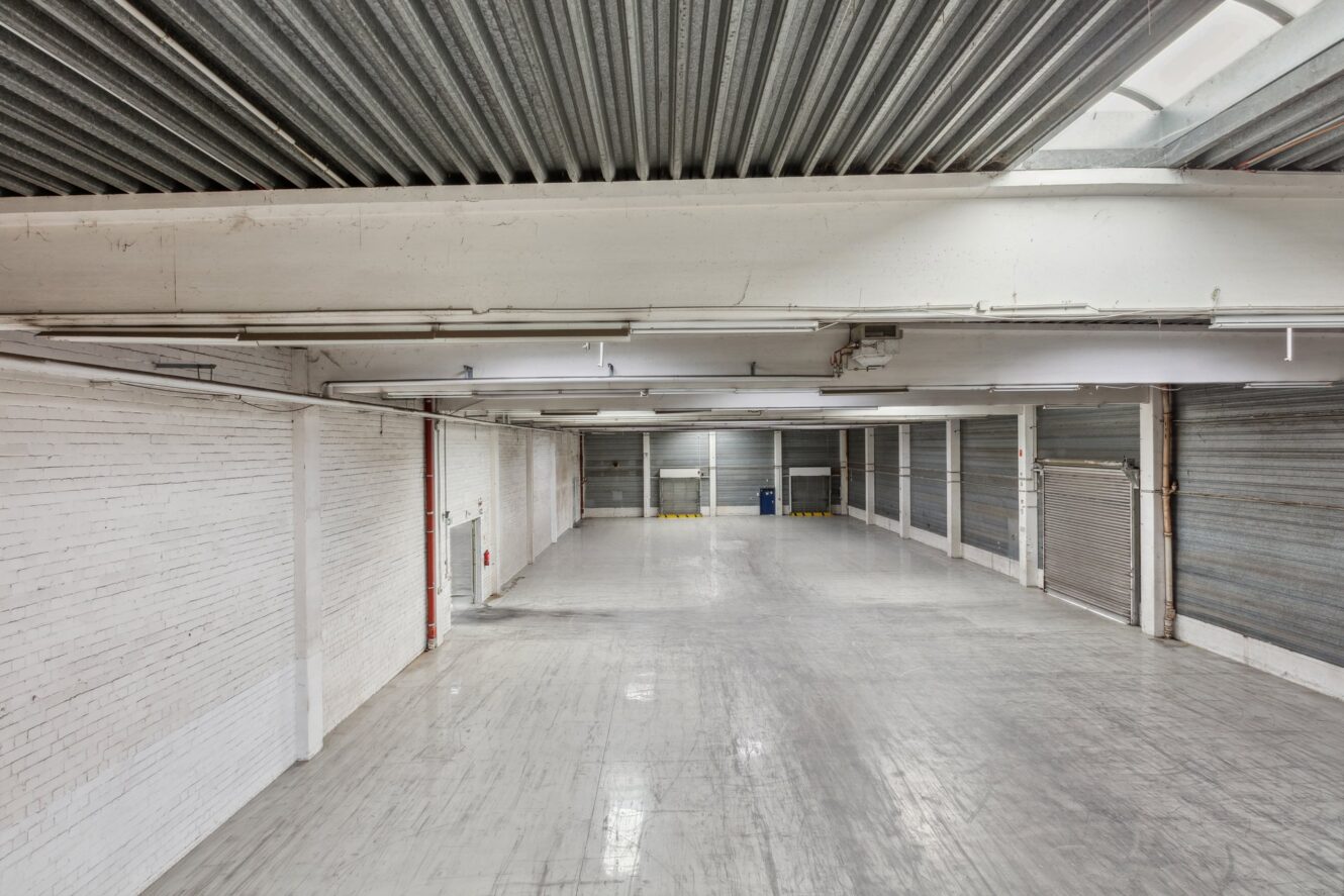 Leeres Industrielager mit weißen Ziegelwänden, Betonboden, Metalldecke und mehreren geschlossenen Ladetoren an einer Seite.