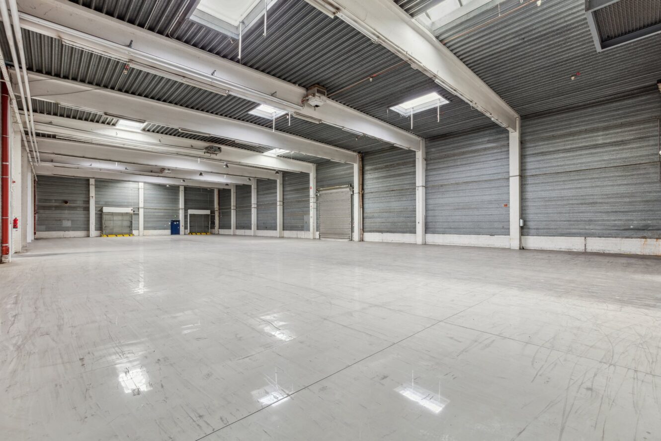 Großes, leeres Industrielager mit polierten Böden, hohen Decken, Metallwänden und mehreren Oberlichtern. Oberlichter und geschlossene Laderaumtore sind sichtbar.