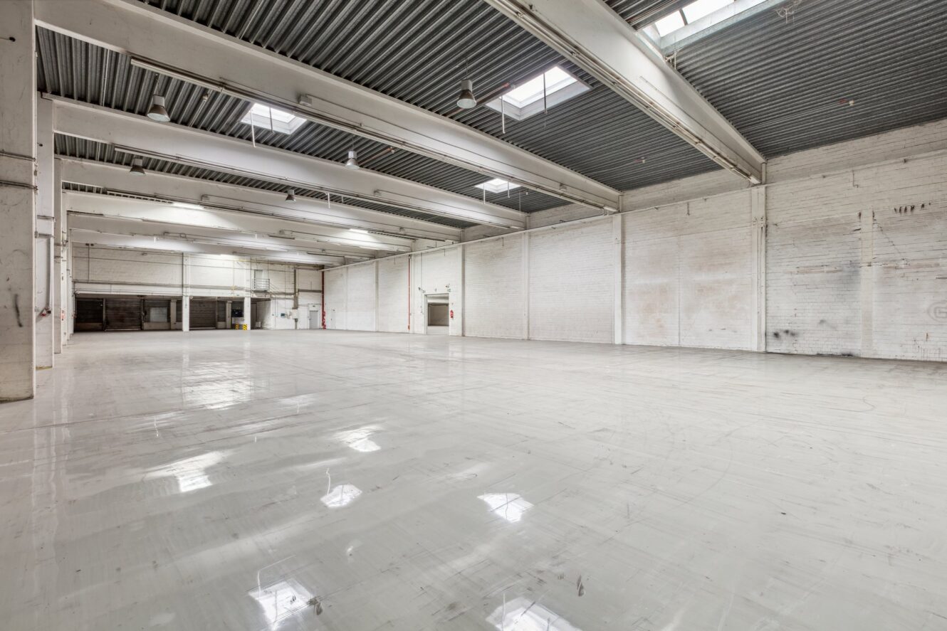 Großes, leeres Industrielager mit weiß gestrichenen Ziegelwänden, hohen Decken, Oberlichtern und polierten Betonböden.