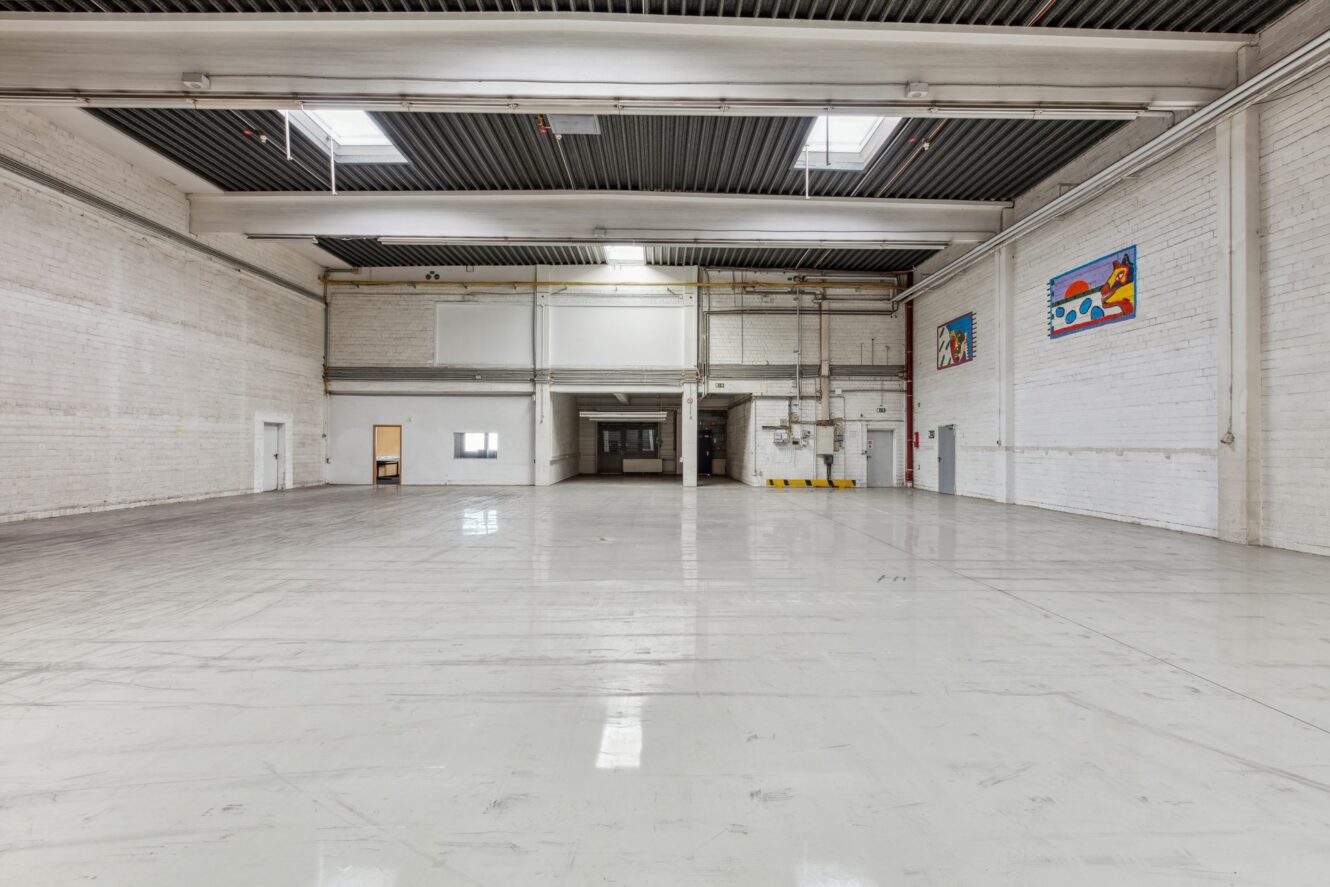 Geräumiges, leeres Industrielager mit weißen Wänden, poliertem Betonboden, hoher Decke, Oberlichtern und bunten Wandmalereien an der rechten oberen Wand.