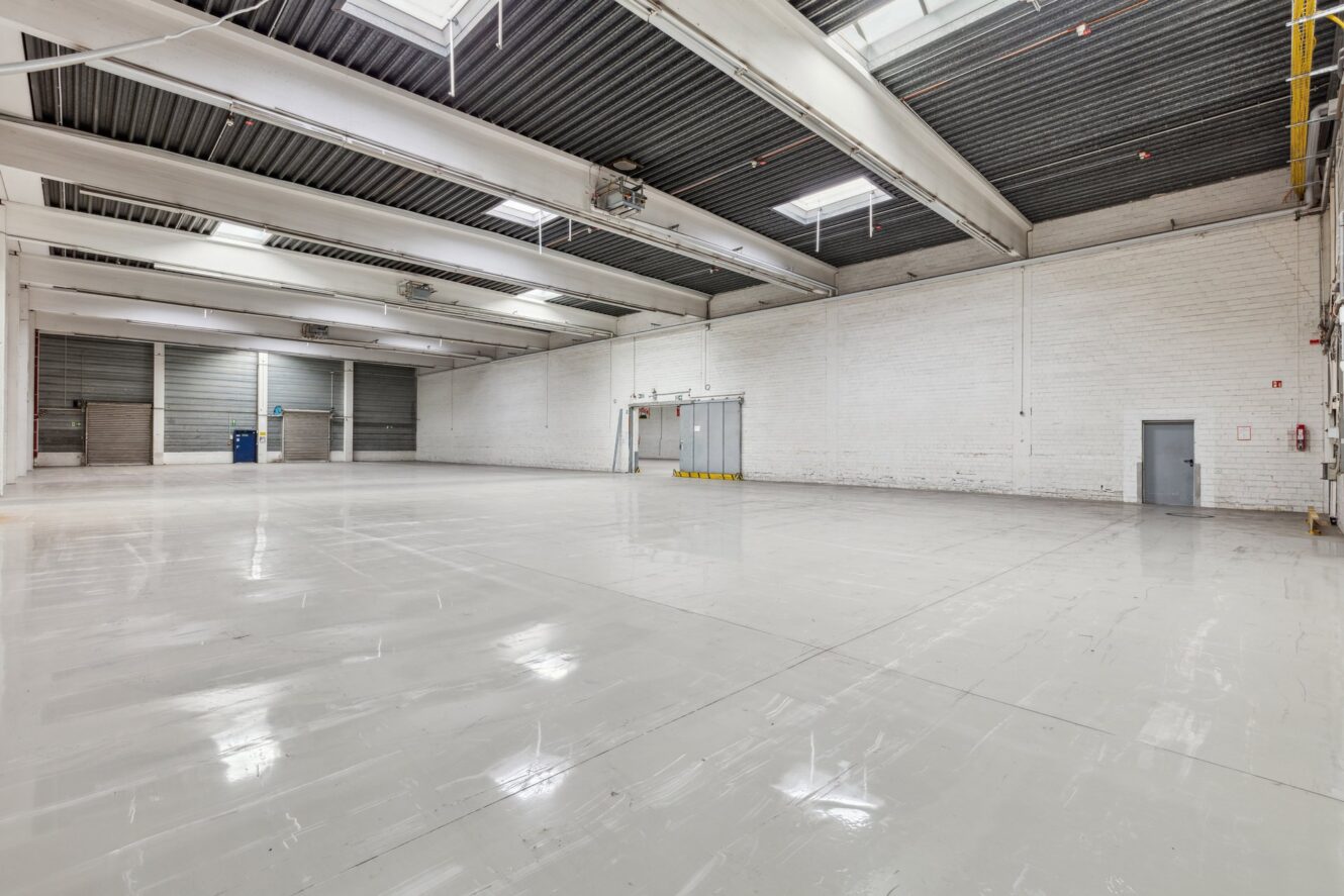 Großes leeres Lagerhaus mit polierten weißen Böden, hohen Decken, freiliegenden Balken, Oberlichtern und weißen Ziegelwänden. An den Wänden sind mehrere Türen zu sehen.