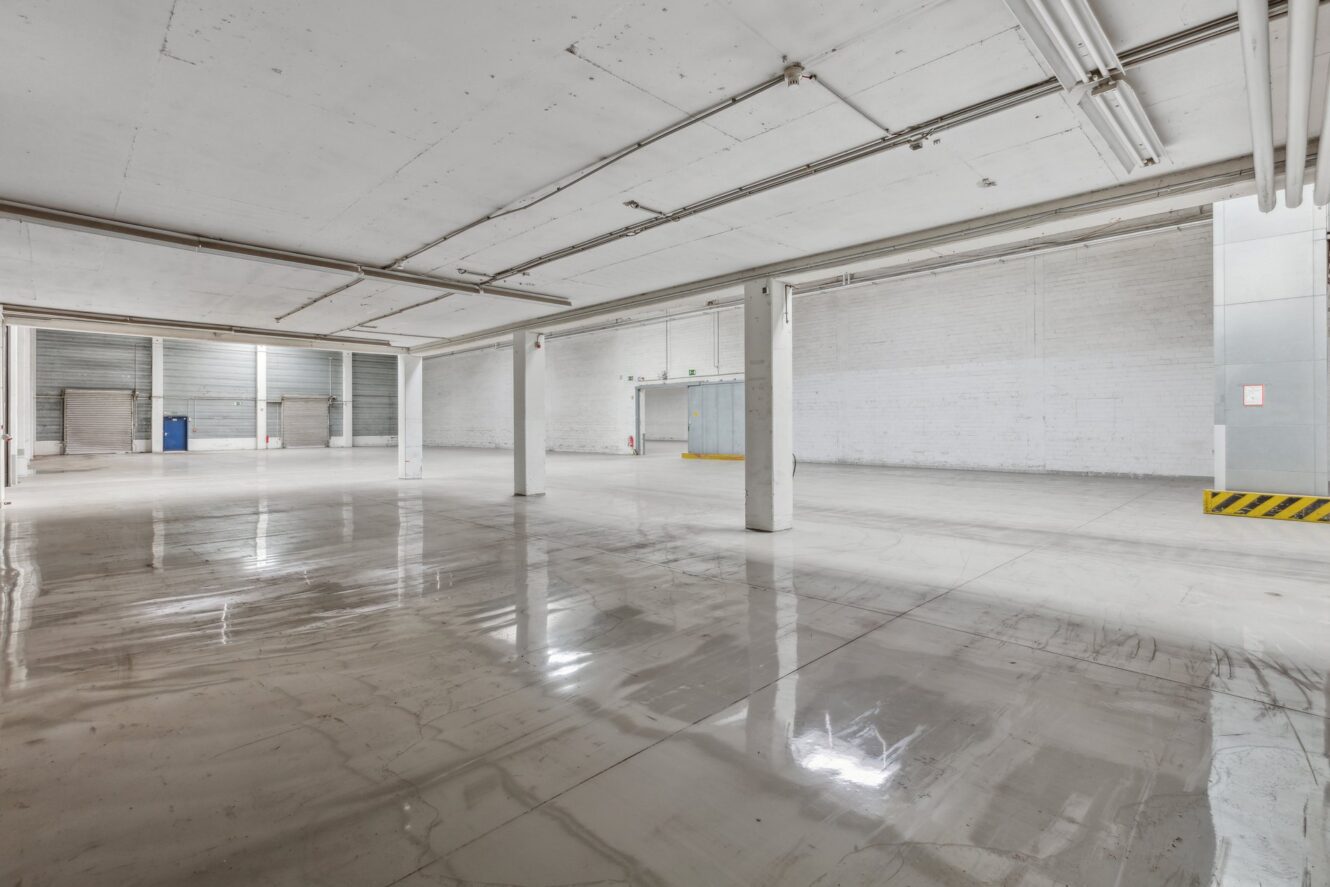 Leeres Industrielager mit polierten Betonböden, weißen Wänden, freiliegenden Rohren und großen Rolltoren an der hinteren Wand.