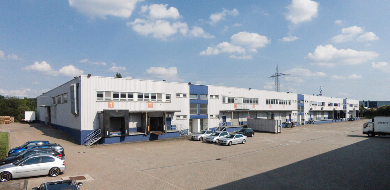 Großes industrielles Lagergebäude mit mehreren Laderampen, geparkten Autos, Lastwagen und einem klaren Himmel mit vereinzelten Wolken.